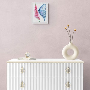 May include: A white dresser with three drawers and gold handles. The dresser has a white top with a clock and a book on it. A white vase with a hole in the side is on the top of the dresser with dried flowers. A framed watercolor painting of a blue butterfly on a pink flower is on the wall behind the dresser.
