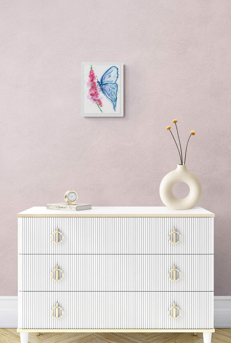 May include: A white dresser with three drawers and gold handles. The dresser has a white top with a clock and a book on it. A white vase with a hole in the side is on the top of the dresser with dried flowers. A framed watercolor painting of a blue butterfly on a pink flower is on the wall behind the dresser.