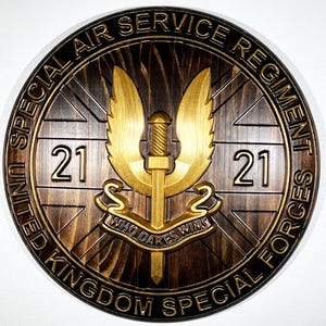 Op de afbeelding: Ronde houten plaquette met de tekst "UNITED SPECIAL AIR SERVICE REGIMENT FORCES UNITED KINGDOM SPECIAL FORCES". Het midden toont een goudkleurige gevleugelde dolk en de tekst "WHO DARES WINS". Het nummer "21" staat aan weerszijden.