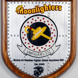 May include: A wooden plaque with a silver metal shield featuring a yellow airplane with a red and white umbrella, surrounded by red dots. The text "Moonlighters" is in yellow letters on a black background. The text "VMFA(AW)-332" is in white letters on a black background. The text "Marine All Weather Fighter Attack Squadron 332 Est. 1943" is in silver letters on a black background. The plaque has a wooden frame with a brown stain.