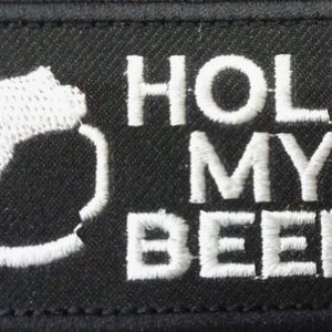 May include: Black embroidered patch with white text that reads "HOLD MY BEER". The patch features a stylized white silhouette of a person's head.