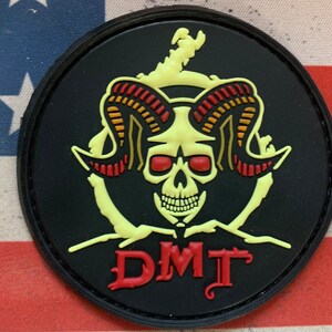 May include: A black round patch with a yellow skull with red eyes and horns. The horns are red, orange, and yellow. The skull is surrounded by a yellow circle. The text "DMT" is written in red below the skull.