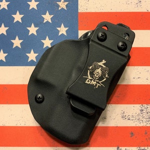 May include: Black holster with a metal clip featuring a skull and ram horns design, with the letters "DMT" below. The holster is set against a backdrop of the American flag.