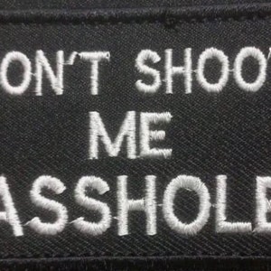 May include: Black rectangular patch with white embroidered text. The text reads "DON'T SHOOT ME ASSHOLE". The patch has a black border and is likely designed to be sewn onto clothing or bags.
