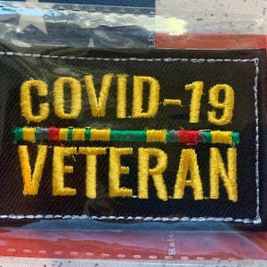 May include: A black embroidered patch with white stitching. The patch says "COVID-19 VETERAN" in yellow lettering with a green, red, and gray stripe above the word "VETERAN".