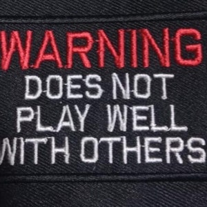 May include: A black patch with white and red embroidered text that reads "WARNING DOES NOT PLAY WELL WITH OTHERS".