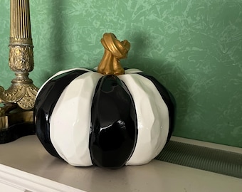 Op de afbeelding: Een decoratieve pompoen met afwisselende zwarte en witte verticale strepen. De pompoen heeft een goudkleurige steel en staat op een wit oppervlak. De pompoen is een herfstthema decoratie.