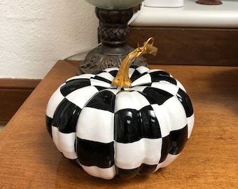 Op de afbeelding: Een decoratieve pompoen met een zwart-wit geblokt patroon. De pompoen heeft een goudkleurige steel en staat op een houten oppervlak. Een herfstthema item voor huisdecoratie.