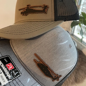 May include: Two trucker hats with a brown leather helicopter patch. The top hat is olive green with a black mesh back. The bottom hat is gray with a black mesh back. The bottom hat has a Richardson brand label with the text "EST. 1972" and "TRUCKER".
