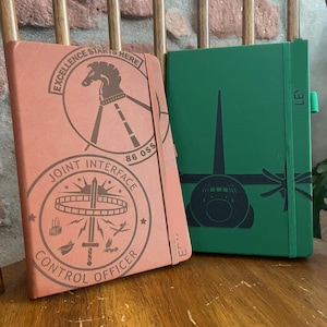 May include: A copper-colored notebook with two circular stamps featuring a horse head and text reading EXCELLENCE STARTS HERE 86 OSS and JOINT INTERFACE CONTROL OFFICER, next to a green notebook with a dark graphic of a cargo aircraft and text.