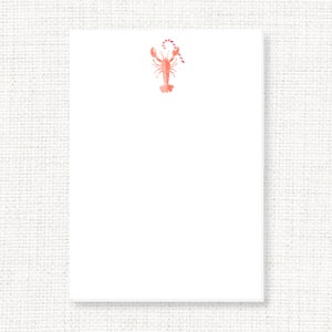 May include: A white notepad with a coral-colored lobster illustration holding a candy cane at the top. The lobster and candy cane are in a watercolor style. The notepad is rectangular and has a clean, minimalist design.