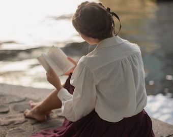 May include: A person wearing a white blouse and a burgundy skirt sits by the water, reading a book. A picnic blanket with croissants, strawberries, and coffee is spread out nearby. The scene is set under a stone bridge.