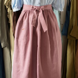 May include: A light blue blouse with puffed sleeves and a pink apron. The apron has a pocket and a bow at the waist. The apron is made of a soft, textured fabric. Clothing items hang in the background.