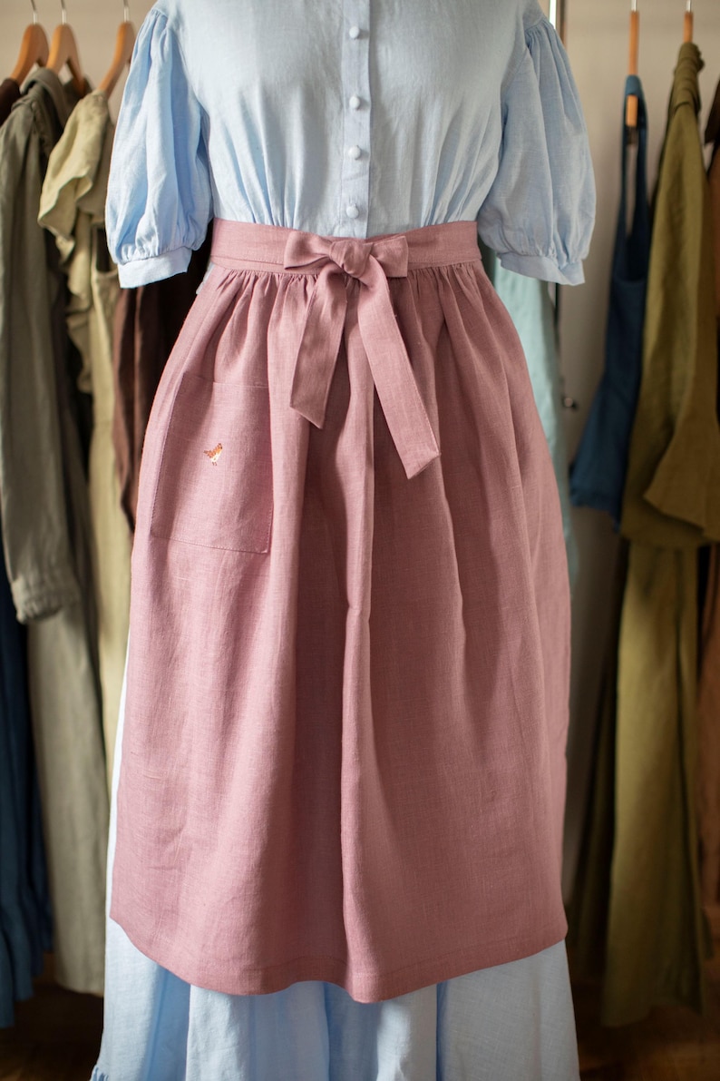 May include: A light blue blouse with puffed sleeves and a pink apron. The apron has a pocket and a bow at the waist. The apron is made of a soft, textured fabric. Clothing items hang in the background.