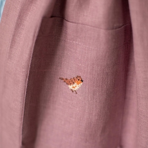 May include: Close-up of a dusty rose-colored linen garment with a pocket. A small, embroidered bird with brown, orange, and white details is on the pocket. The fabric has a textured appearance.