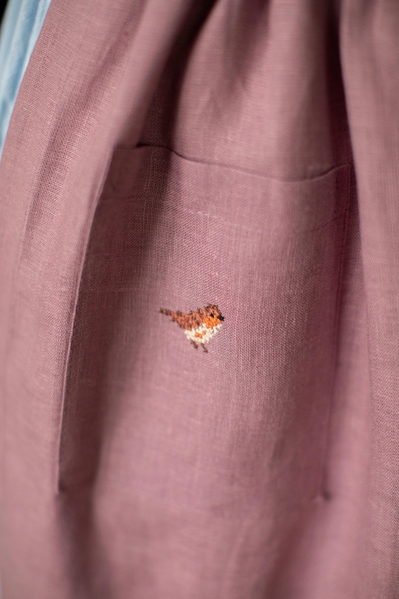 May include: Close-up of a dusty rose-colored linen garment with a pocket. A small, embroidered bird with brown, orange, and white details is on the pocket. The fabric has a textured appearance.
