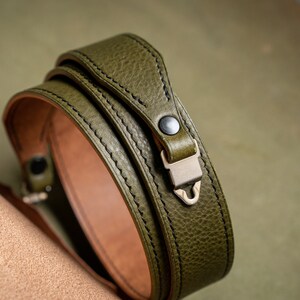 May include: A green leather camera strap with a metal clasp. The strap is folded in half and the clasp is visible.