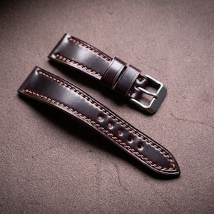May include: Two brown leather watch straps with orange stitching. The strap on the left has a buckle attached. The straps are on a brown surface.