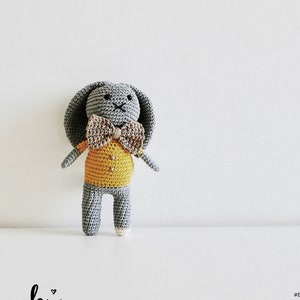 Puede incluir: Un conejito de crochet gris que lleva una camisa amarilla y una pajarita marrón. El conejito se llama Benjamin Conejo. La imagen incluye el texto "bechi handmade" y "#bechihandmade".