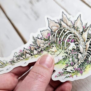 Pode incluir: Um adesivo branco com uma ilustra&ccedil;&atilde;o detalhada de um esqueleto de dinossauro coberto de flores cor-de-rosa e folhagem verde. O adesivo est&aacute; sobre uma superf&iacute;cie de madeira branca.