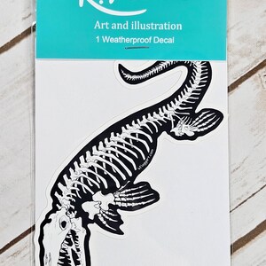 May include: A black and white sticker featuring a skeleton of a prehistoric marine reptile. The sticker is packaged in a clear plastic sleeve with a turquoise background and the text "K. FLAGG Art and illustration 1 Weatherproof Decal".