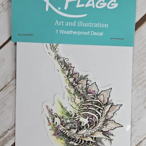 Pode incluir: Um adesivo &agrave; prova de intemp&eacute;ries que apresenta uma ilustra&ccedil;&atilde;o detalhada de um esqueleto de dinossauro com flores e vegeta&ccedil;&atilde;o crescendo de seus ossos. O adesivo &eacute; branco com contornos pretos e acentos rosa, verdes e marrons. O texto "K.FLAGG Art and illustration 1 Weatherproof Decal" est&aacute; impresso em um fundo turquesa.
