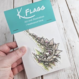 Pode incluir: Um decalque &agrave; prova de intemp&eacute;ries que apresenta uma ilustra&ccedil;&atilde;o detalhada de um drag&atilde;o esquel&eacute;tico com flores crescendo de seus ossos. O decalque &eacute; embalado em um saco de pl&aacute;stico transparente com um cabe&ccedil;alho turquesa que diz "K. FLAGG Art and Illustration 1 Weatherproof Decal".