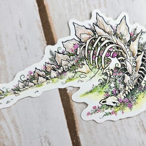 Pode incluir: Um adesivo branco com uma ilustra&ccedil;&atilde;o detalhada de um esqueleto de dinossauro cercado por flores cor-de-rosa e folhagem verde. O dinossauro est&aacute; deitado de lado com a cabe&ccedil;a apoiada no ch&atilde;o. O adesivo tem um fundo branco e um contorno preto.