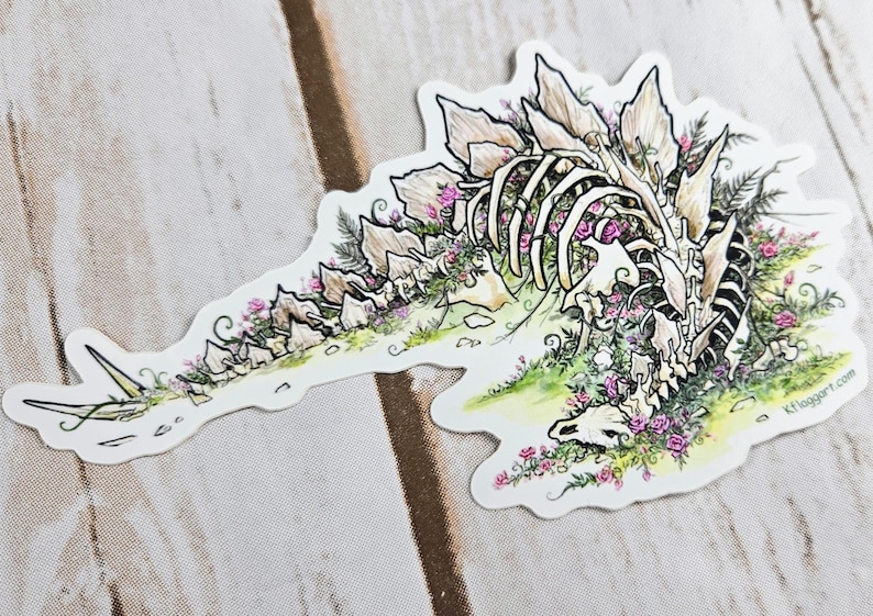 Pode incluir: Um adesivo branco com uma ilustra&ccedil;&atilde;o detalhada de um esqueleto de dinossauro cercado por flores cor-de-rosa e folhagem verde. O dinossauro est&aacute; deitado de lado com a cabe&ccedil;a apoiada no ch&atilde;o. O adesivo tem um fundo branco e um contorno preto.