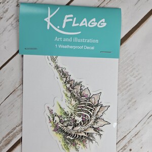 Pode incluir: Um decalque &agrave; prova de intemp&eacute;ries com uma ilustra&ccedil;&atilde;o detalhada de um esqueleto rodeado de flores e folhagem. O decalque &eacute; embalado em uma manga de pl&aacute;stico transparente com um cabe&ccedil;alho turquesa que diz "K. FLAGG Art and illustration 1 Weatherproof Decal".