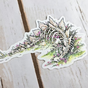 Pode incluir: Um adesivo branco com uma ilustra&ccedil;&atilde;o detalhada de um esqueleto de dinossauro coberto de flores cor-de-rosa. O dinossauro est&aacute; deitado de lado com a cabe&ccedil;a apoiada no ch&atilde;o. O adesivo tem um fundo branco e o texto "K1 Swagger.com" no canto inferior direito.