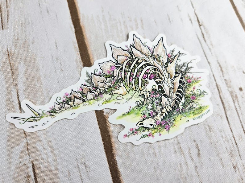 Pode incluir: Um adesivo branco com uma ilustra&ccedil;&atilde;o detalhada de um esqueleto de dinossauro coberto de flores cor-de-rosa. O dinossauro est&aacute; deitado de lado com a cabe&ccedil;a apoiada no ch&atilde;o. O adesivo tem um fundo branco e o texto "K1 Swagger.com" no canto inferior direito.