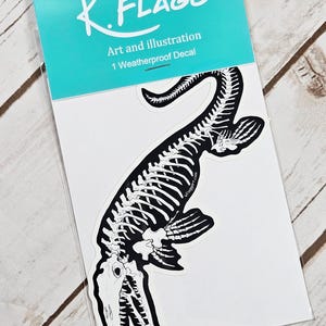 May include: A black and white sticker featuring a detailed illustration of a plesiosaur skeleton. The sticker is packaged in a clear plastic bag with a turquoise label that reads "K.FLAGG Art and illustration 1 Weatherproof Decal".