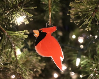 May include: A red cardinal Christmas ornament with a black head and beak. The ornament is hanging on a Christmas tree with white lights.