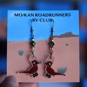 May include: A pair of earrings featuring a cartoon roadrunner design with a brown body and black eyes. The earrings are on a card that reads "MO/KAN ROADRUNNERS RV CLUB".