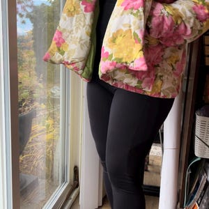 Puede incluir: Una chaqueta acolchada de estampado floral rosa y amarillo con cuello chal. La chaqueta se lleva sobre un top negro sin mangas y leggings negros. La persona lleva calcetines de colores con estampados.
