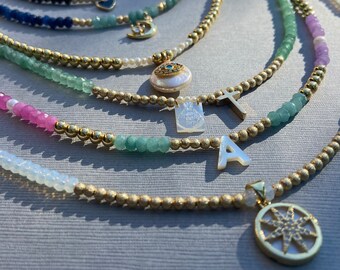 May include: Layered necklaces with gold beads and various charms, including a crescent moon, an evil eye, a cross, a letter A, and a starburst. The necklaces are made with green, pink, and white beads.