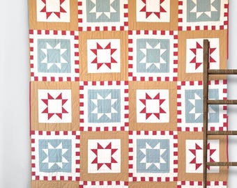 May include: A large, handmade quilt with a repeating pattern of squares. Each square features a star design in red or light blue against a white background, framed by a red border. The quilt is displayed against a wall.