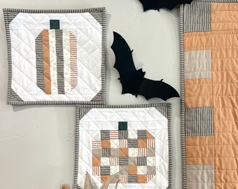Peut inclure: Trois tentures murales matelassées sur le thème de la citrouille. Les tentures sont faites avec un fond blanc et présentent différents motifs de tissu orange, brun et blanc. Les tentures sont exposées sur un mur blanc avec un bouquet de fleurs séchées dans un vase en dessous.
