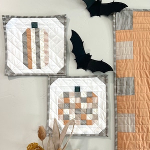 Peut inclure: Trois tentures murales matelassées sur le thème de la citrouille. Les tentures sont faites avec un fond blanc et présentent différents motifs de tissu orange, brun et blanc. Les tentures sont exposées sur un mur blanc avec un bouquet de fleurs séchées dans un vase en dessous.