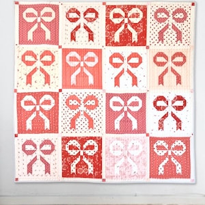 May include: A handmade quilt featuring a repeating pattern of red and white bows. The quilt is composed of sixteen squares, each showcasing a different bow design. The bows are set against various backgrounds, including solid colors and polka dots. The quilt is hanging on a white wall.