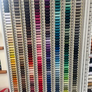 Puede incluir: Un expositor de pared de hilo Gutermann Creativ Sew-all en carretes de 100 m. Los carretes están dispuestos en filas de diferentes colores, incluyendo beige, marrón, amarillo, naranja, rojo, rosa, morado, azul, verde y gris.