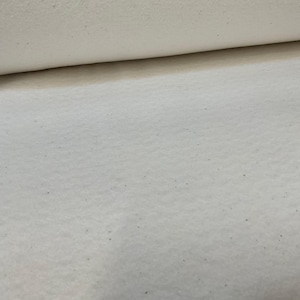 May include: Close-up of a roll of off-white fabric. The fabric has a slightly textured surface with small, dark flecks throughout. The top edge of the roll is visible, showing the rolled layers of the material. The fabric appears soft and possibly natural.