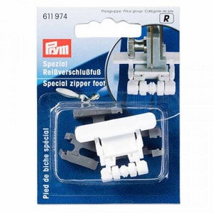 Puede incluir: Un pie de cremallera de plástico blanco para máquinas de coser. El pie está empaquetado en una tarjeta azul y blanca con el texto "Spezial Reißverschlußfuß Special zipper foot" y el número "611 974".