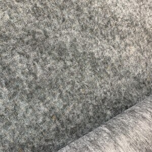 Puede incluir: Primer plano de una tela de fieltro gris con una superficie texturizada. El material tiene un aspecto moteado con variaciones de color, que van del gris claro al gris oscuro. La tela está enrollada en la esquina inferior derecha.