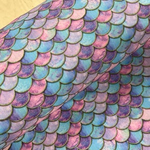 May include: A roll of fabric featuring a mermaid scale pattern. The design incorporates overlapping, semi-circular shapes in shades of blue, purple, and pink, with a subtle gold outline. The fabric has a textured appearance.