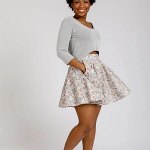 May include: A woman wearing a gray, long-sleeved crop top and a flared skirt with a colorful floral pattern. The skirt has pockets. She is wearing pink sneakers.