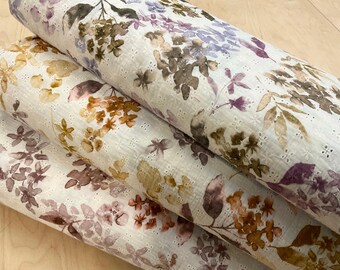 May include: Close-up of several rolls of white eyelet fabric with a floral pattern. The fabric features watercolor-style flowers in shades of purple, brown, and gold. The eyelet design adds texture and visual interest to the fabric.