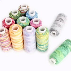 Könnte beinhalten: Eine Sammlung bunter Garnrollen, jede mit einem weißen Kern. Die Garne sind in verschiedenen Farben erhältlich, darunter Rosa, Gelb, Grün, Blau und Grau. Die Rollen sind auf einer weißen Oberfläche angeordnet, ideal für Näh- und Bastelprojekte.