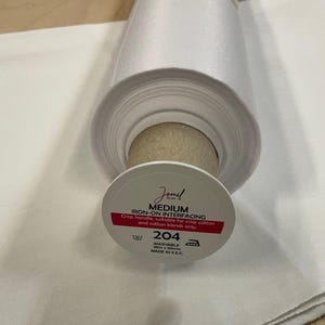Pode incluir: Um rolo de tecido de entretela termocolante branco com uma etiqueta que diz "Jomel MEDIUM IRON-ON INTERFACING". A etiqueta também indica que o tecido é lavável e adequado para misturas de algodão. O rolo tem 25 metros x 90 cm.